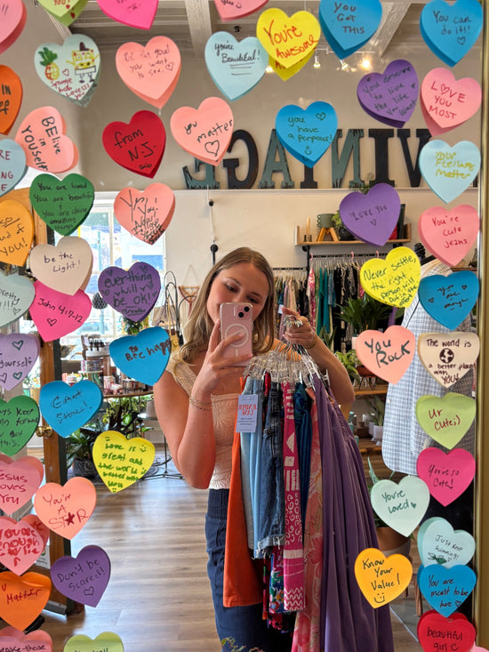 Person standing in front of a decorative mirror with colorful heart-shaped stickers, holding vintage clothing on consignment in Staunton Virginia juniper lane vintage boutique things to do and shopping